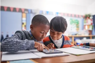 Two young boys in a classroom look at a tablet together, with books and another tablet on the desk.