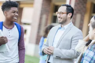 A smiling teacher talks with students outside a school building on a sunny day.