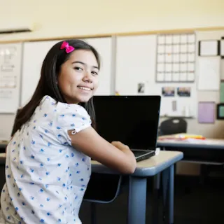 Smiling girl with a pink hair clip sits at a desk with a laptop in a classroom setting.