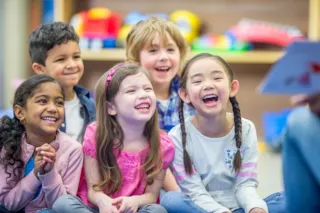Children sitting in a group, smiling and laughing, while listening to a person reading a book in a classroom setting.