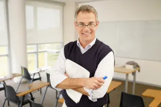 Smiling man with glasses holding papers and a pen, standing in an empty classroom with desks and a whiteboard.