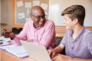 Teacher and student smiling and talking at a desk with a laptop and open binder in a classroom.
