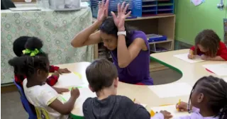 A teacher playfully makes antlers with her hands while young children draw at a classroom table.