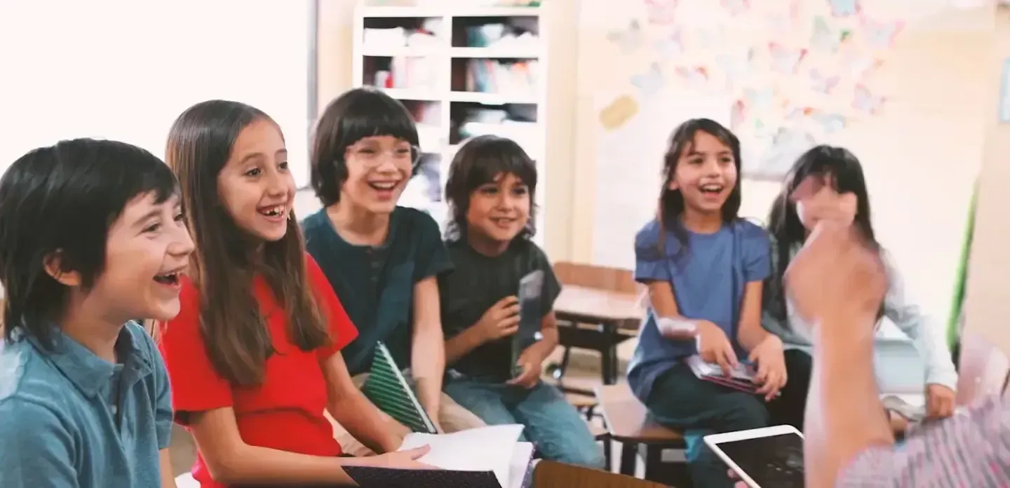 Six children sit in a classroom, smiling and laughing together while looking at a teacher.