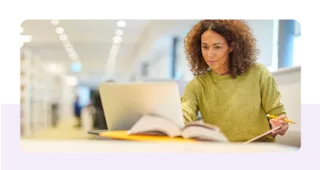 Woman with curly hair working on a laptop at a desk, holding a pen. An open book is in the foreground, inside a library.