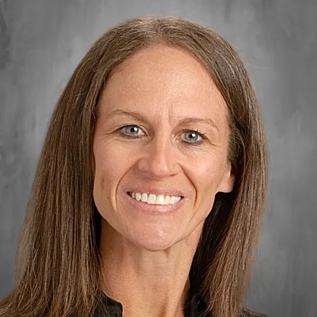 A woman with short curly brown hair smiles at the camera against a gray background.