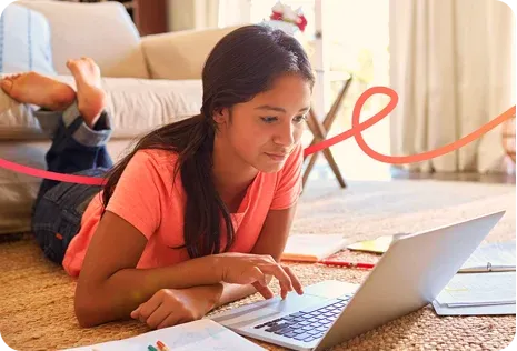 A girl lies on the floor, using a laptop and surrounded by notebooks and papers in a cozy living room.