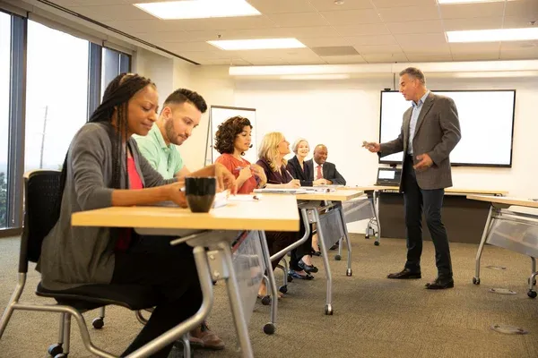 A man stands and speaks to a group of people seated at desks in a modern conference room.