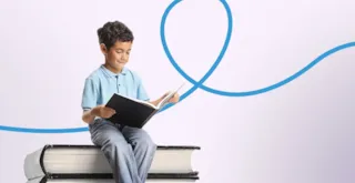 A young boy sitting on a stack of oversized books and reading a book.