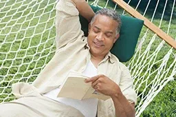 A man reclines in a hammock reading a book in a summer setting with a relaxed expression on his face
