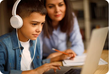 A boy wearing headphones uses a laptop while a woman sits beside him, assisting with schoolwork.