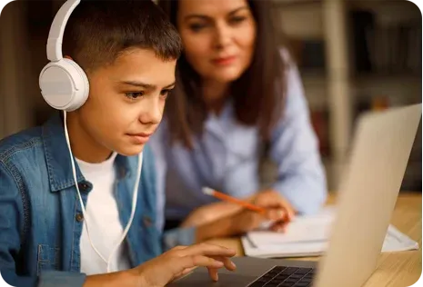 A boy wearing headphones uses a laptop while a woman sits beside him, assisting with schoolwork.
