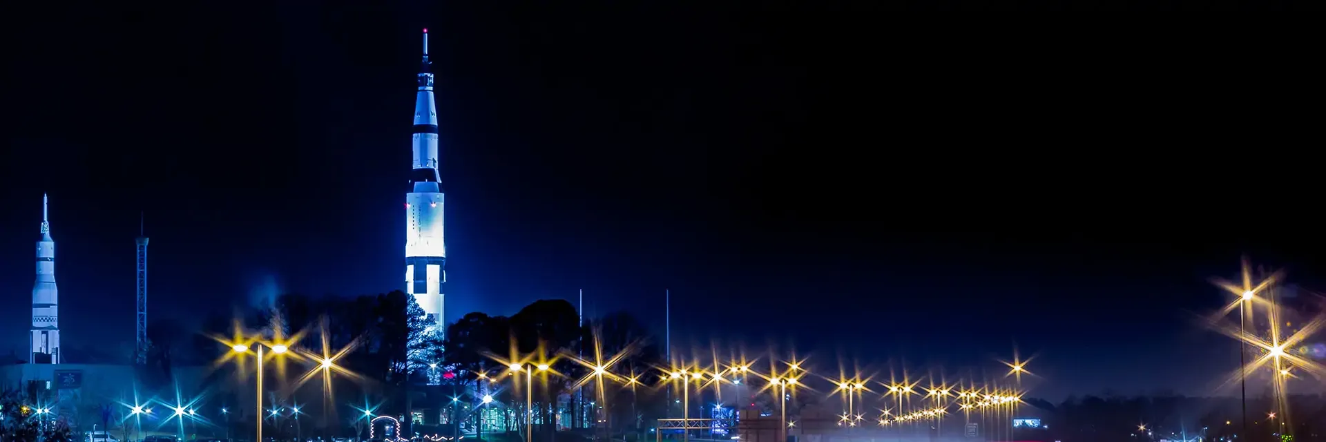 A tall skyscraper illuminated at night, surrounded by trees and streetlights, against a dark blue sky.