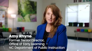 A woman in a blue blazer sits in a classroom; text shows her name and title: Amy Rhyne, Former Senior Director.