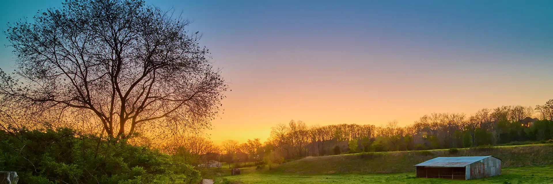 A barn on a grassy field at sunrise, with trees in the background and a clear, colorful sky.