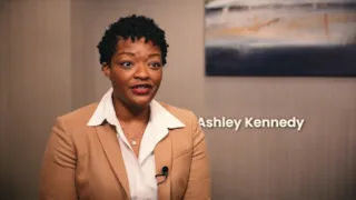 Woman in a tan blazer, white shirt, and microphone lavalier, speaking. Name Ashley Kennedy appears beside her. Abstract art in background.