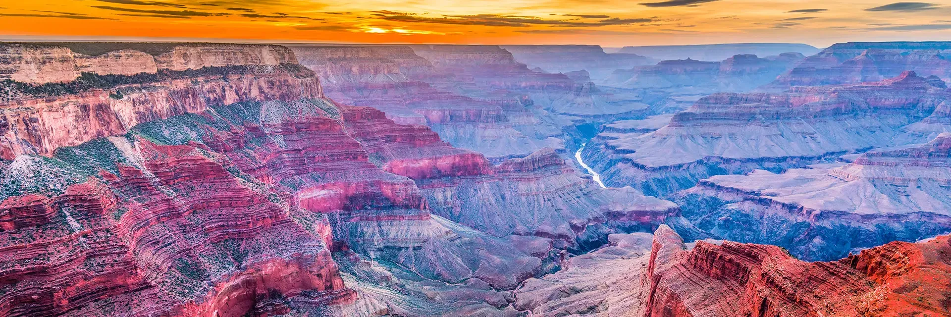 Grand Canyon at sunset with colorful rock layers, deep valleys, and sunlight illuminating the horizon.