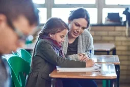 A teacher assists a young student with her work at a desk in a classroom, with another student blurred in the foreground.