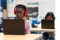 Two students in a classroom use laptops. The student in the foreground wears headphones, focused on the screen.