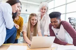 A group of students and a teacher smile while looking at a laptop together in a bright classroom.