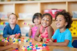 Four young children sit at a table, smiling and playing with colorful interlocking toys in a classroom setting.