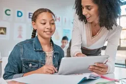 A teacher helps a student with a tablet in a classroom. Theyre smiling, with alphabet posters visible on the walls.