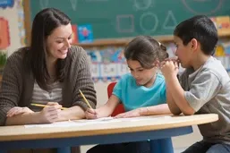 A teacher helps two young students with a writing assignment at a classroom table.