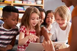 Children sitting together, engaged and smiling, looking at a book in a classroom setting.