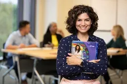 Smiling teacher watches a young boy using a digital tablet in a colorful classroom setting.