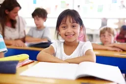 Young girl smiles at her desk in a classroom, with an open notebook in front of her. Other children and a teacher are in the background.