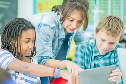 A teacher assists two students using a tablet in a classroom, with colorful educational materials in the background.