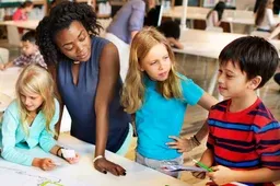 A teacher and three children stand together at a table, talking and working on a classroom project.