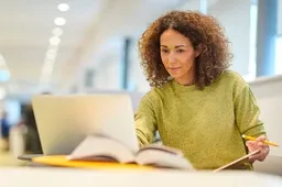 Woman with curly hair working at a desk, using a laptop and holding a pencil with an open book nearby.