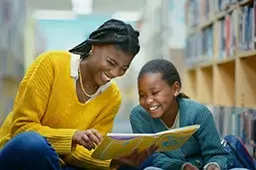 Two girls sit on the floor in a library, smiling and reading a book together.