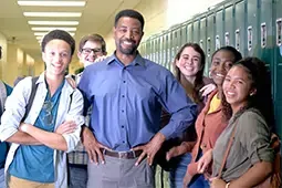 A smiling teacher stands in a hallway with a group of diverse, happy students in front of lockers.