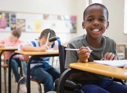 Smiling boy in a wheelchair writing at a desk in a classroom, with two other kids focused on their work in the background.