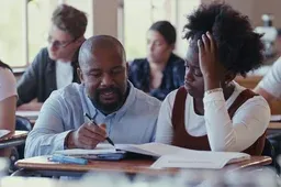 A teacher helps a student with her work at a classroom desk while other students study in the background.
