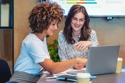 Two women smiling and working together on a laptop at a desk, with papers and coffee cups. A presentation screen is in the background.