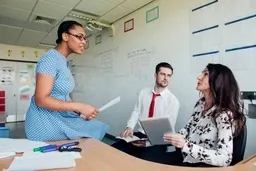 Three people in an office: a woman standing, holding papers, talking to a seated man and woman with a tablet. Whiteboard in background.