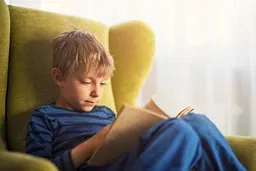 Young boy in blue pajamas sitting in a green armchair, reading a book by a window with soft natural light.