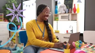 Smiling person in a yellow sweater works on a laptop in a colorful classroom.