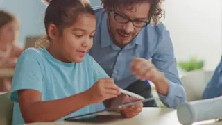 Teacher helping a student use a tablet in a classroom setting, with other students blurred in the background.