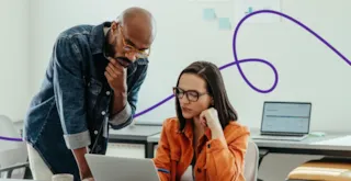 A man and a woman in an office look at a laptop screen together.