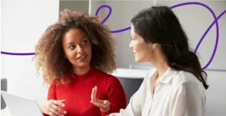 Two women sit together indoors, having a focused conversation near a laptop, with a purple line in the background.