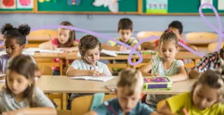 Children sitting at desks in a classroom, writing or drawing, with books and supplies on the tables.