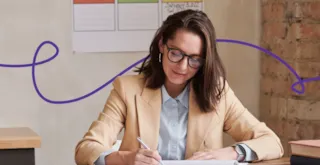 A woman in a tan blazer and glasses writing in a notebook at a desk.