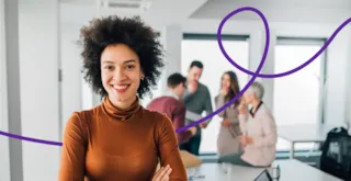 Smiling woman in brown top stands with arms crossed; group of people talk in bright office background.