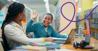 Two students in a library high-five each other while studying at a table with a laptop.