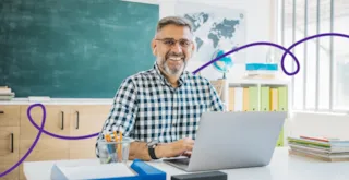 Smiling man with glasses works on a laptop at a desk in a classroom with books and a blackboard.