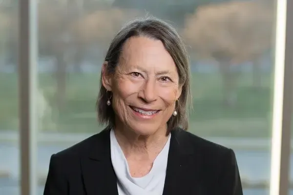 Smiling older woman with gray hair wearing a black blazer and white shirt, standing indoors by large windows.